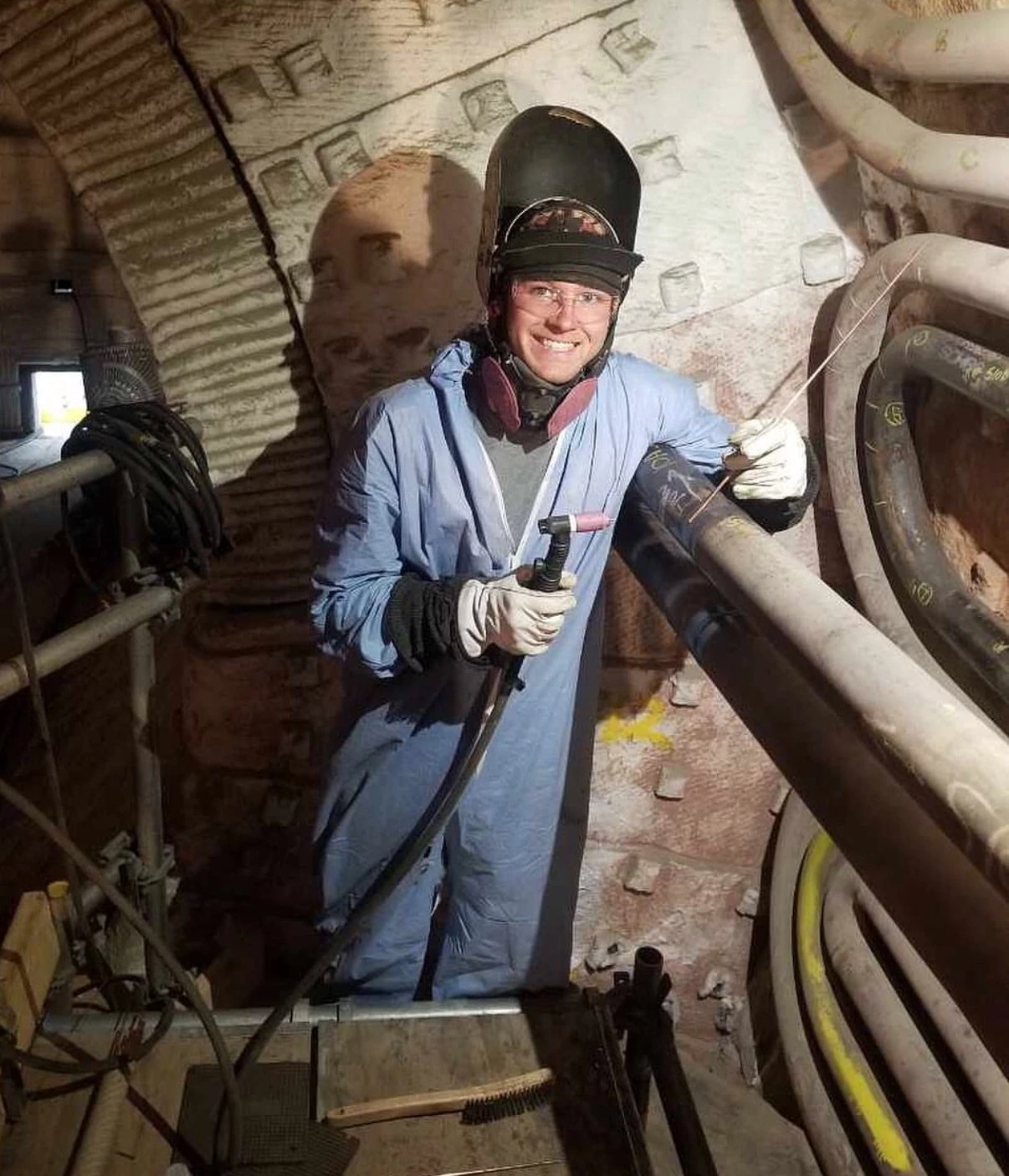 Travis Field with his welding helmet raised and TIG torch beside an industrial pipe assembly